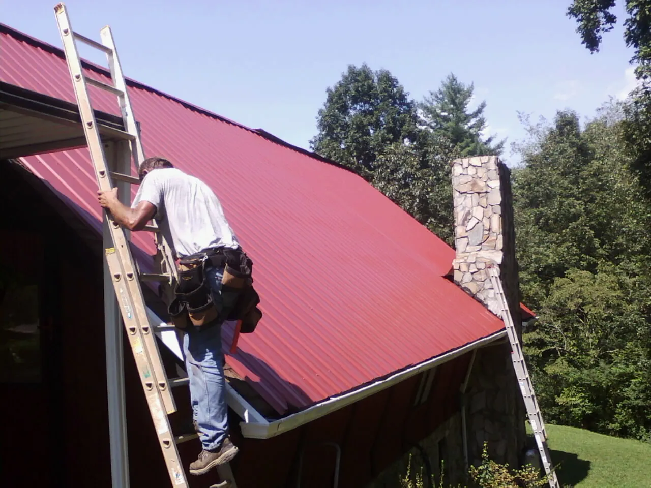 Roofer on a ladder beside a red metal roof and stone chimney — Soffit & Fascia Repair by Roofing Brothers in North Myrtle Beach